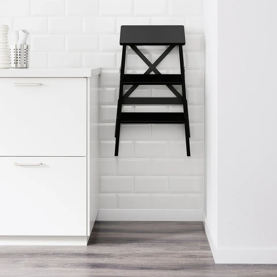 Black folded stepladder leans against white tiled wall. Part of modern minimalist decor.