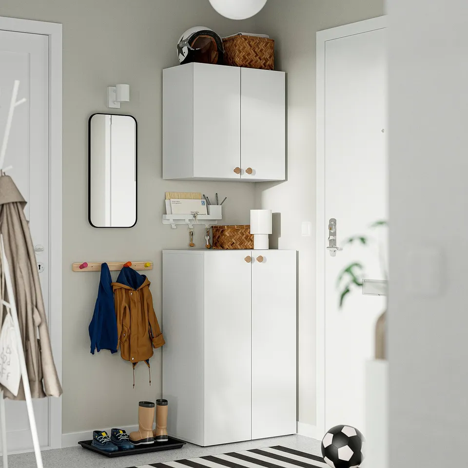A tidy entryway with a white LASTARE storage unit, featuring cabinets and open shelves. Jackets hang beside it, with shoes and a ball nearby.