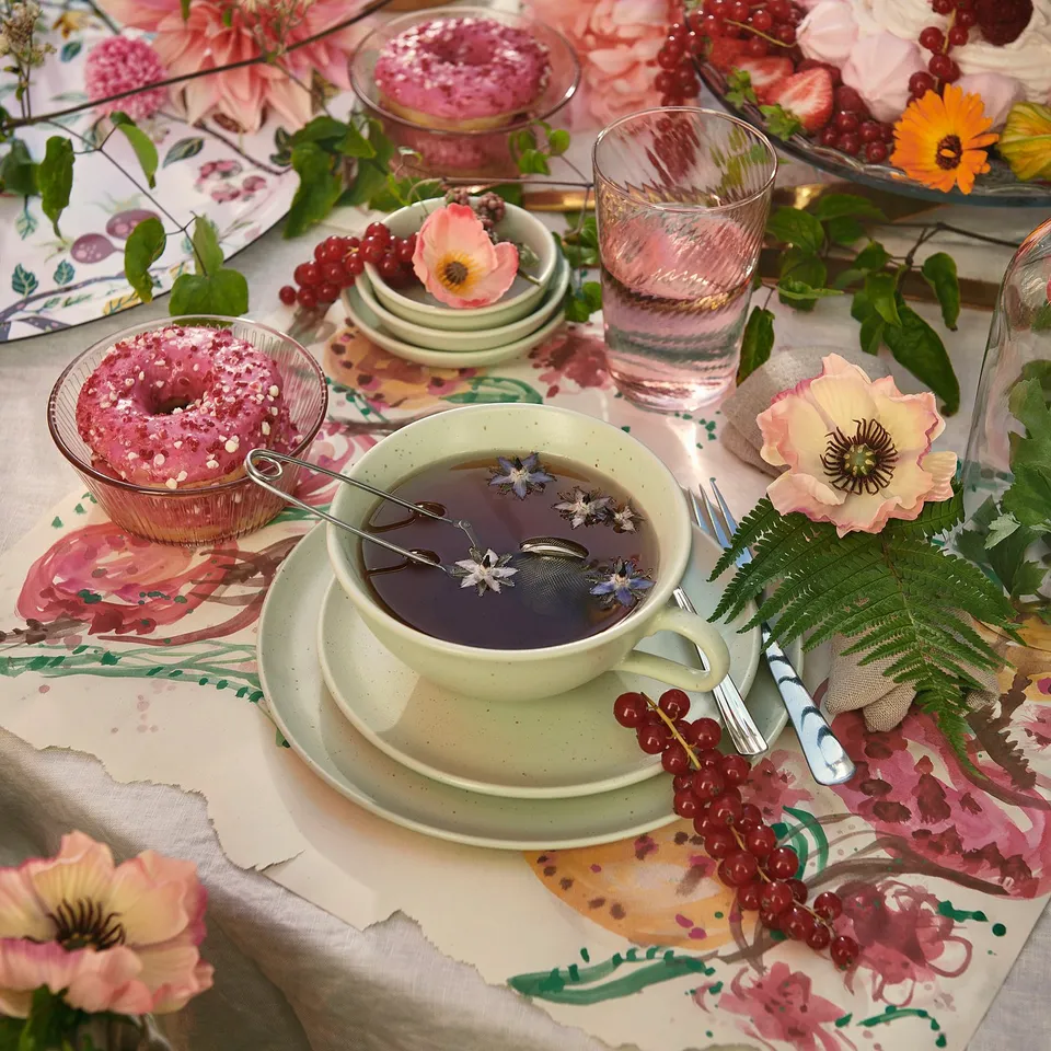 Beautiful table setting with moss green mug and bowls, flowers, and pink pastries on floral tablecloth.