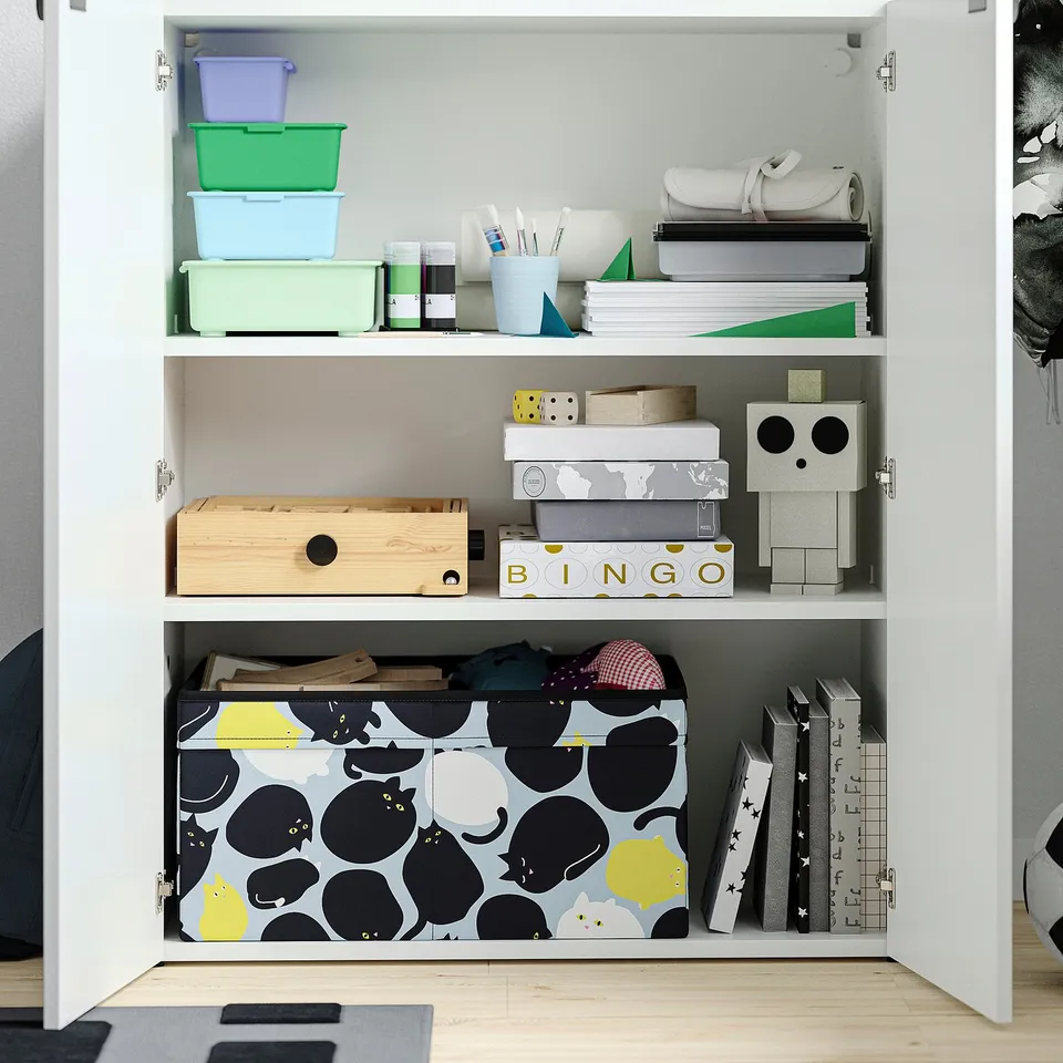 White cupboard with three shelves storing colourful boxes, arts supplies, games, and books; adjustable feet.