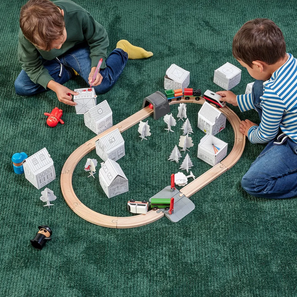 Two children play with a diy cardboard train set, building houses and tracks.