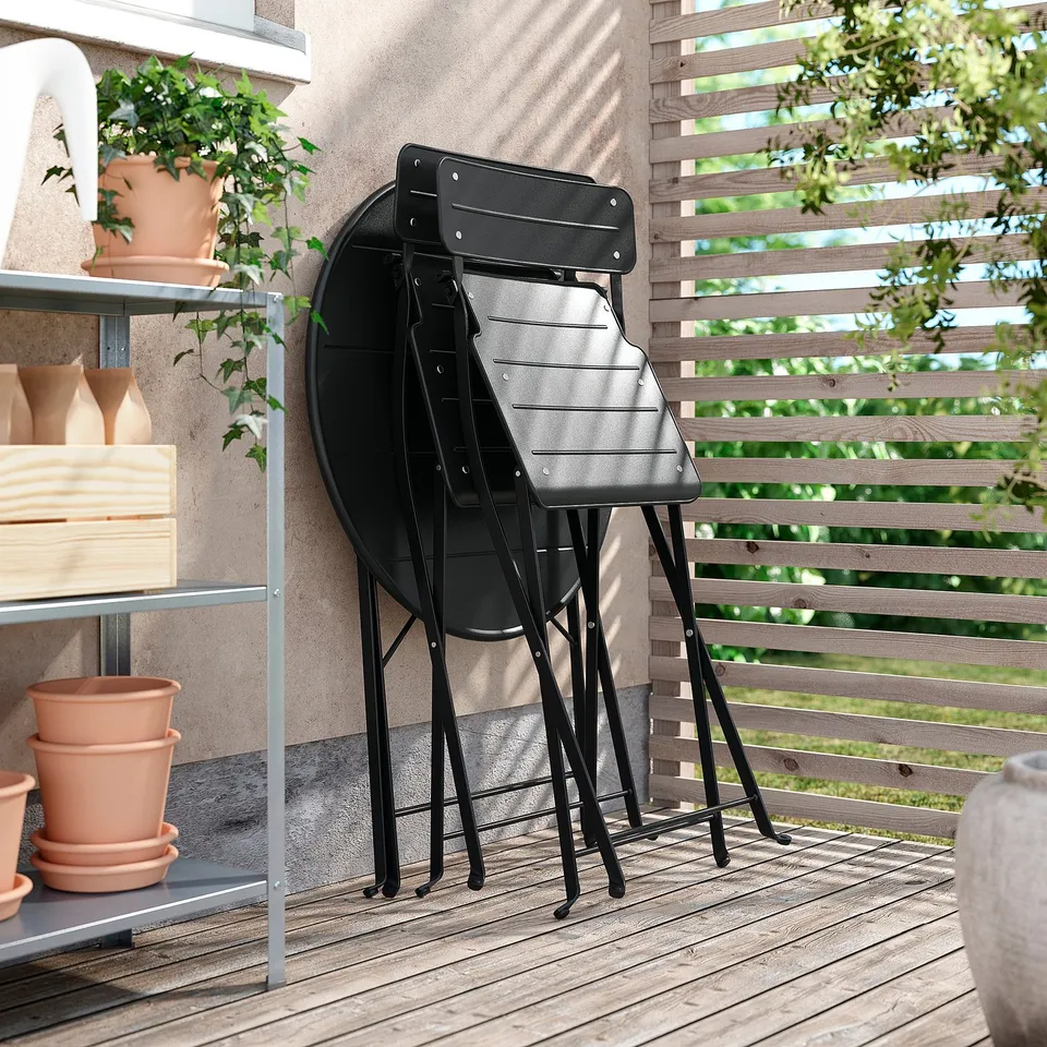 Image 2 black metal SUNDSÖ chairs folded against a wooden panel, ideal for small spaces, placed on a wooden deck near a potted plant.
