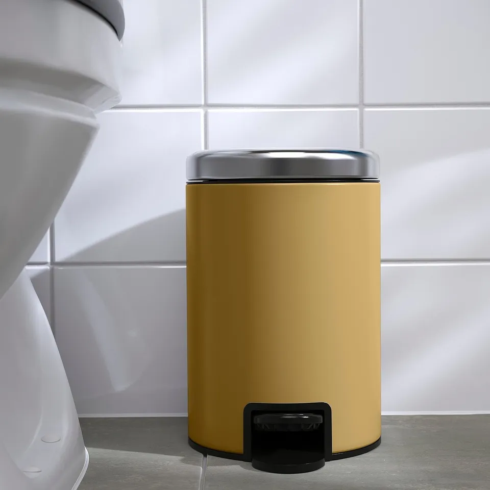 A yellow foot pedalled rubbish can in a bathroom with white walls and a toilet.