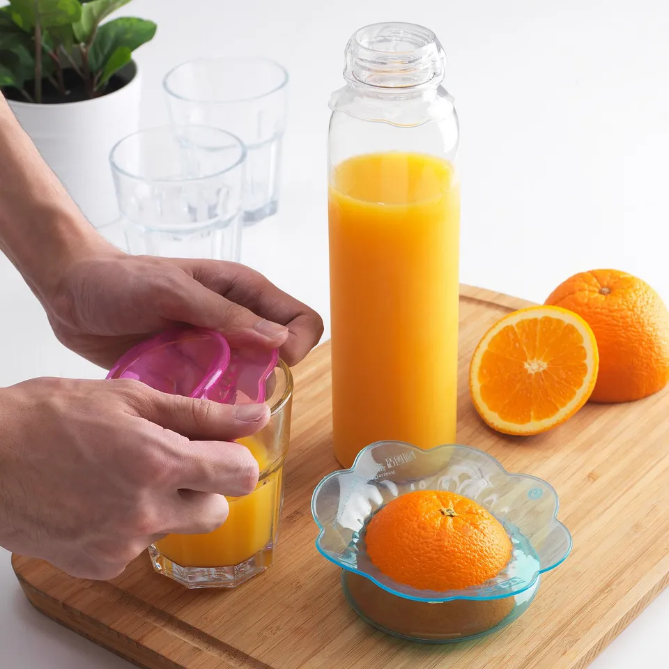 Person covering a glass of orange juice with a reusable food cover. Oranges and other glasses nearby on a wooden board.