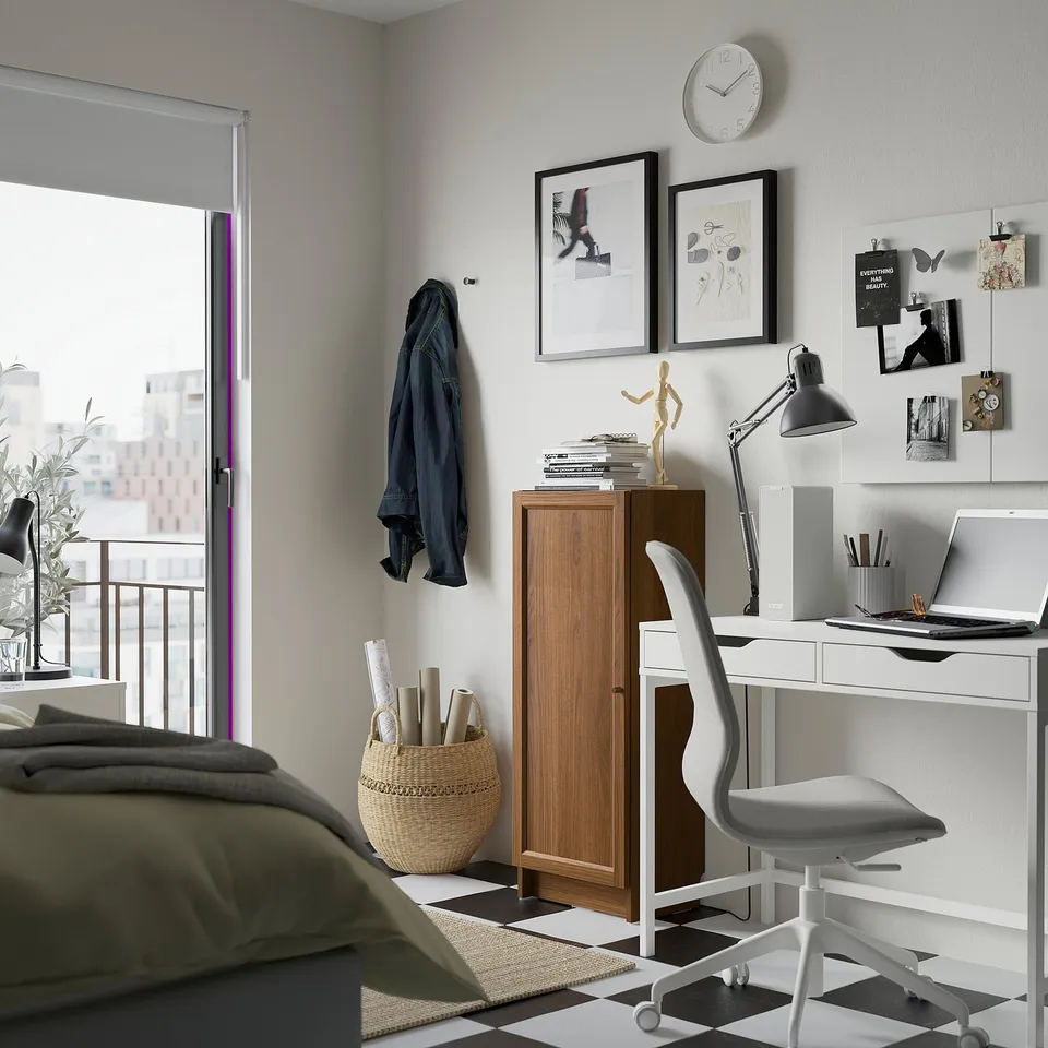 Modern home office: BILLY bookcase, desk with laptop, lamp, clock, jacket, green blanket, bright natural light.
