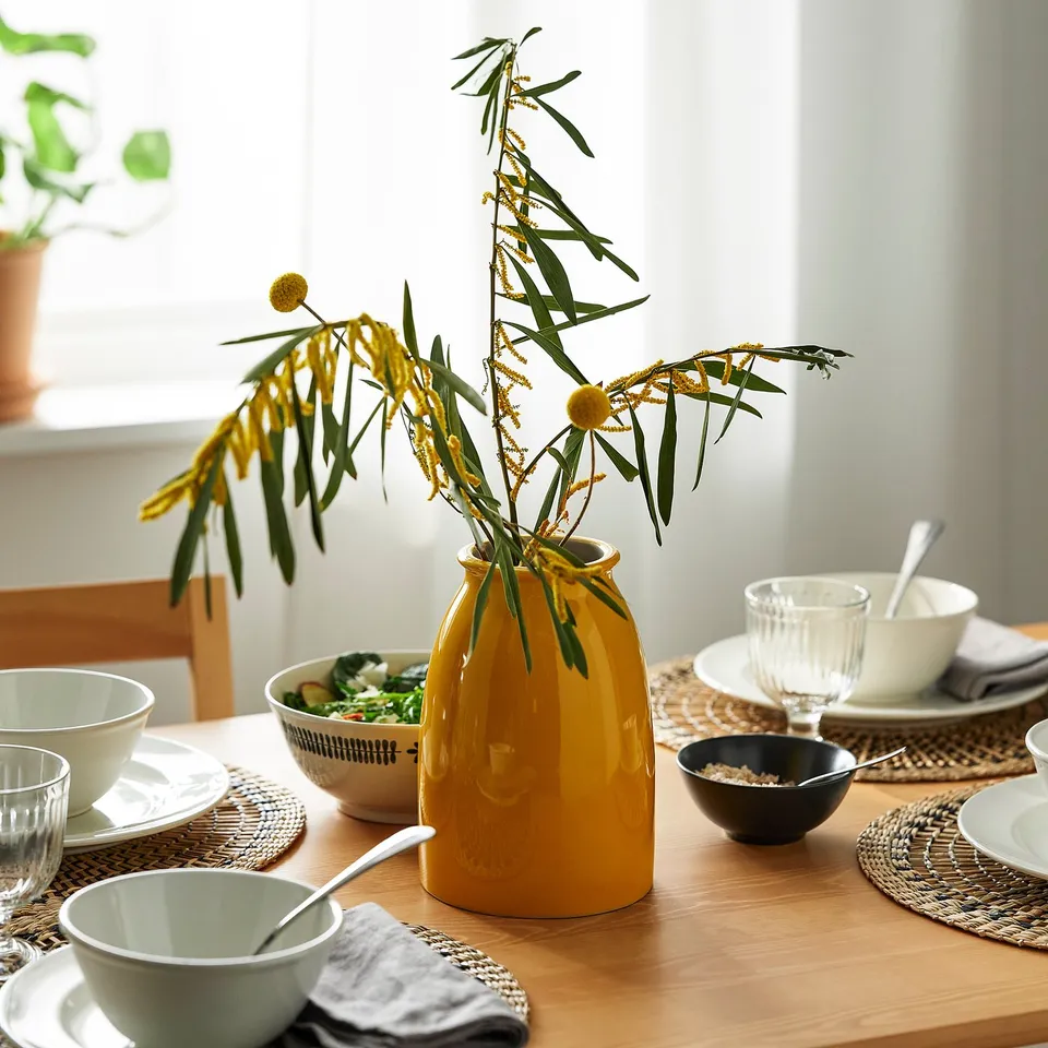 Yellow vase with long yellow flowers on wooden table with bowls and plates. Bright colours, natural feel.