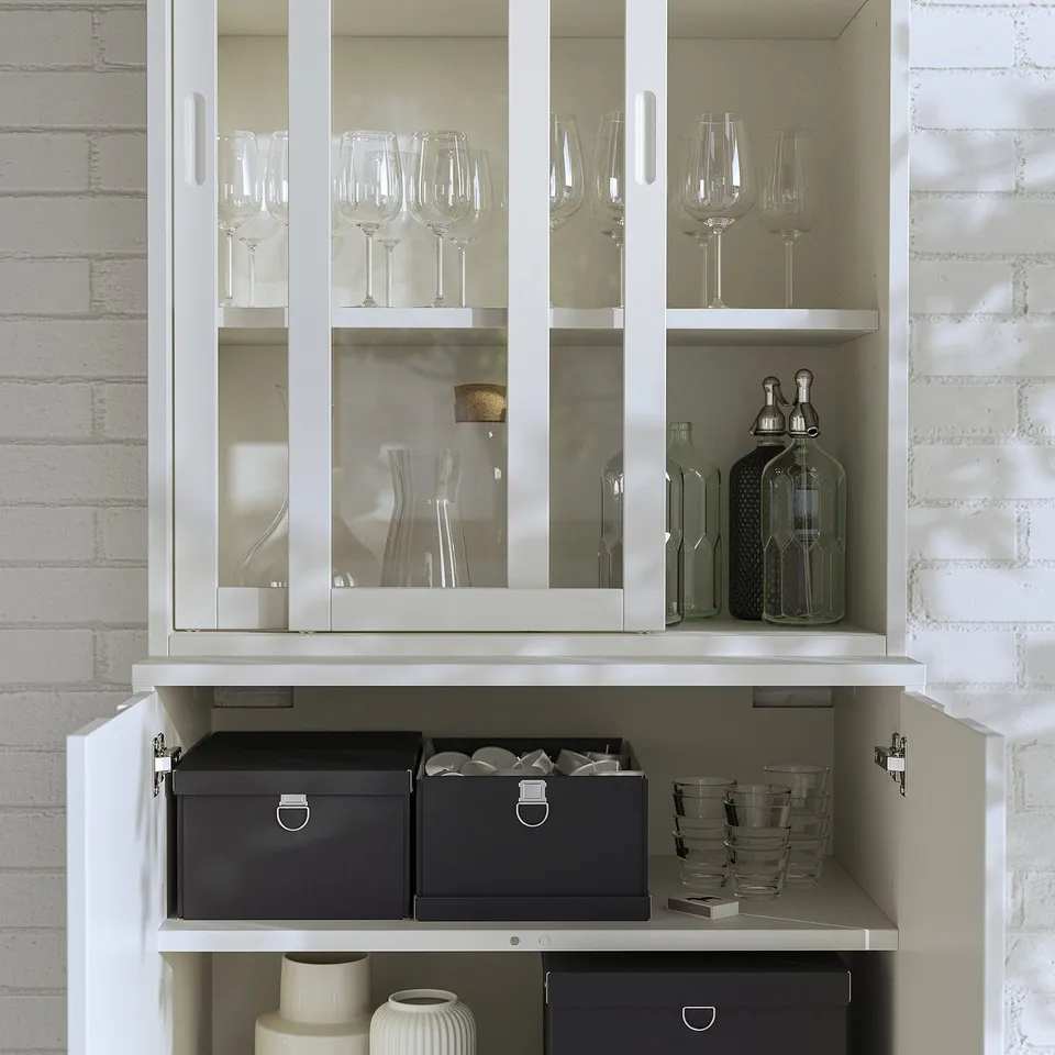 White TONSTAD cabinet with glass doors, storing wine glasses, carafes, grey storage boxes, small cups, and a white vase.