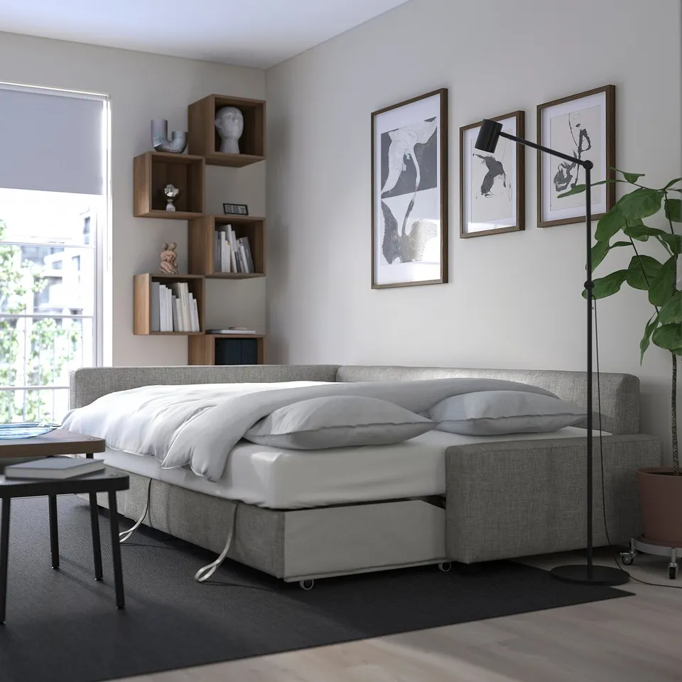 Modern bedroom with FRIHETEN sofa-bed, white bedding, wooden shelves, lamps, and art.