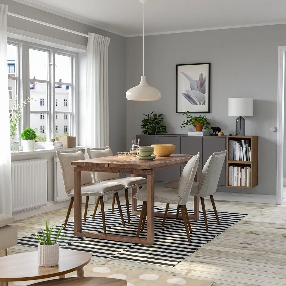 Modern dining set with oak table, fabric chairs, striped rug, dishes, lamp, plants, and decor.