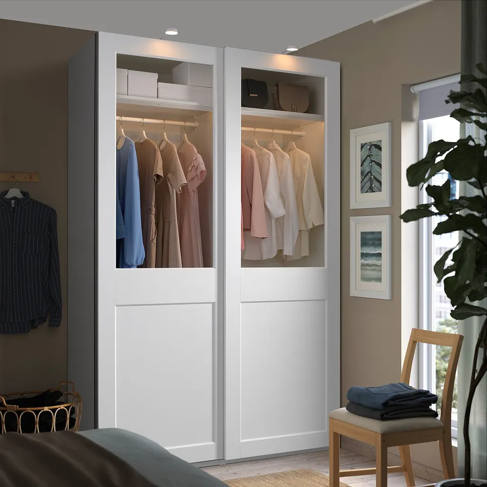 Modern bedroom with PAX wardrobe, sliding doors, one glass, chair with clothes, plant by window, wall art.