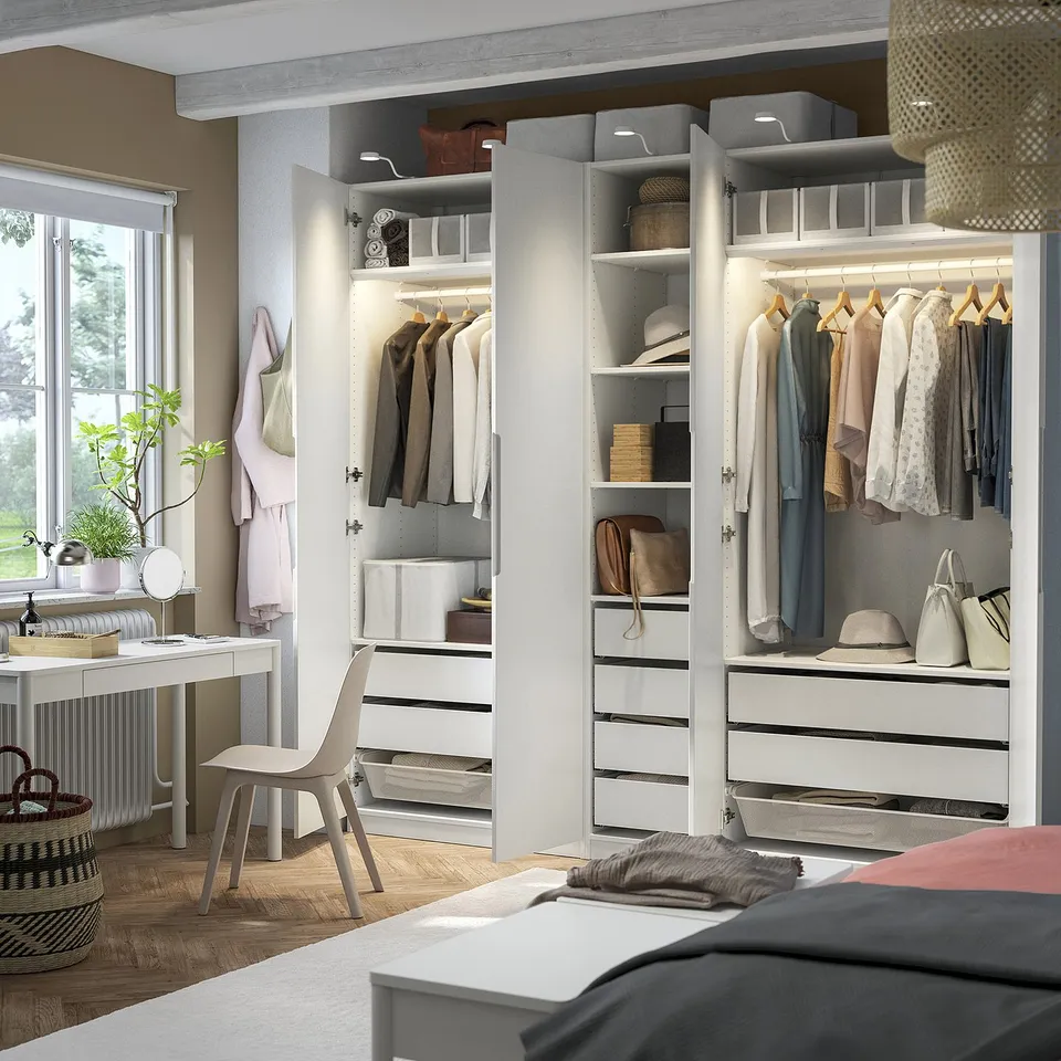 Open PAX wardrobe displaying clothes, shelves, and drawers in a well-lit bedroom.