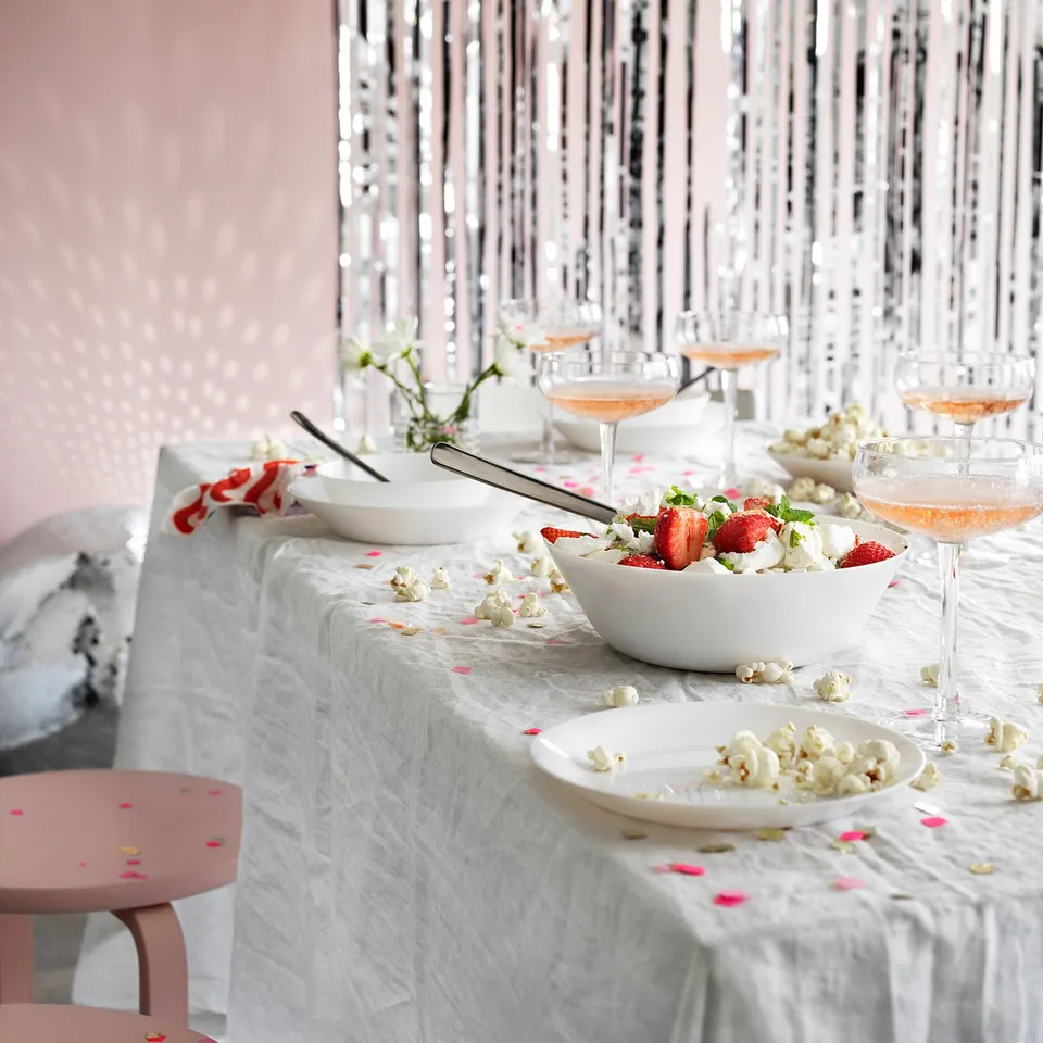 Elegant table setting with white tempered glass bowls, pink confetti, and glasses. Decorative silver streamers in the background.