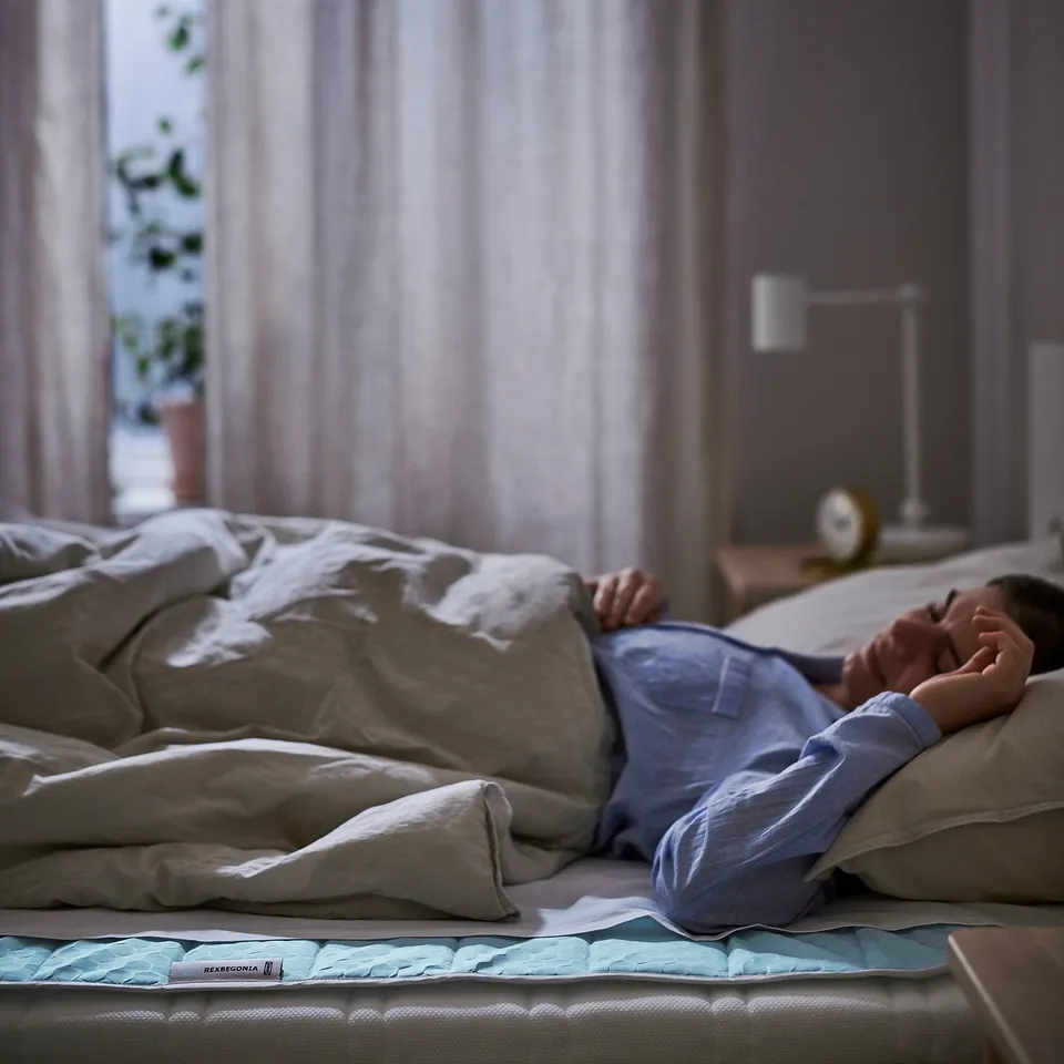 Person in bed using a blue REXBEGONIA mattress protector. Bed has white sheets and pillows, and a clock is on the bedside table.