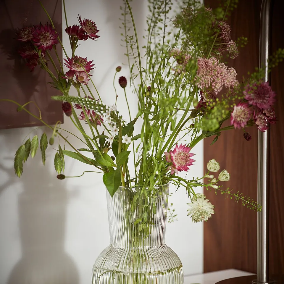 Clear glass vase with vertical lines, holding colourful flowers. Elegant round shape, sits on a white cabinet.