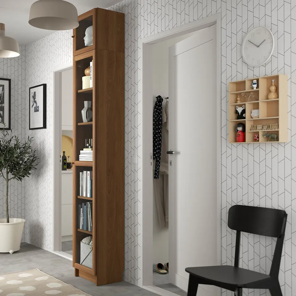 Modern living room with tall BILLY bookcase, black chair, and wall clock. Geometric wallpaper, white door, stylish decor.