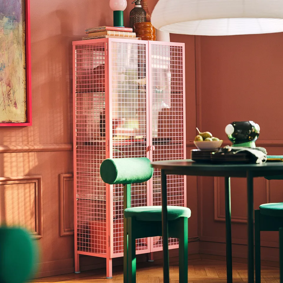 Bright pink metal grid cabinet; sits between wall and table, taller than nearby stools, decorated with books and vases.