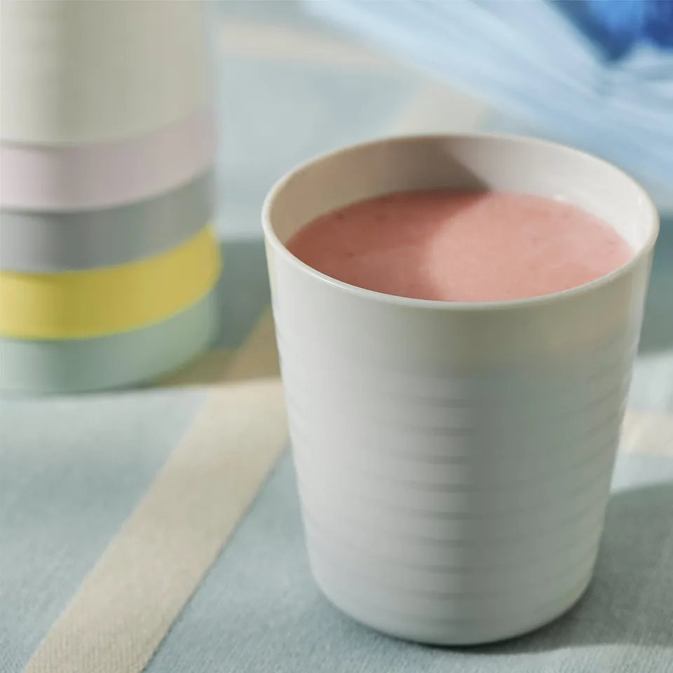 A white plastic cup with horizontal ridges holds pink liquid. Behind it, a colourful stackable cup sits on a light striped tablecloth.