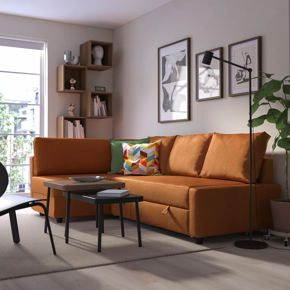 Modern living room with orange sofa-bed, colourful pillows, wooden shelf, floor lamp, coffee table, window, artworks.