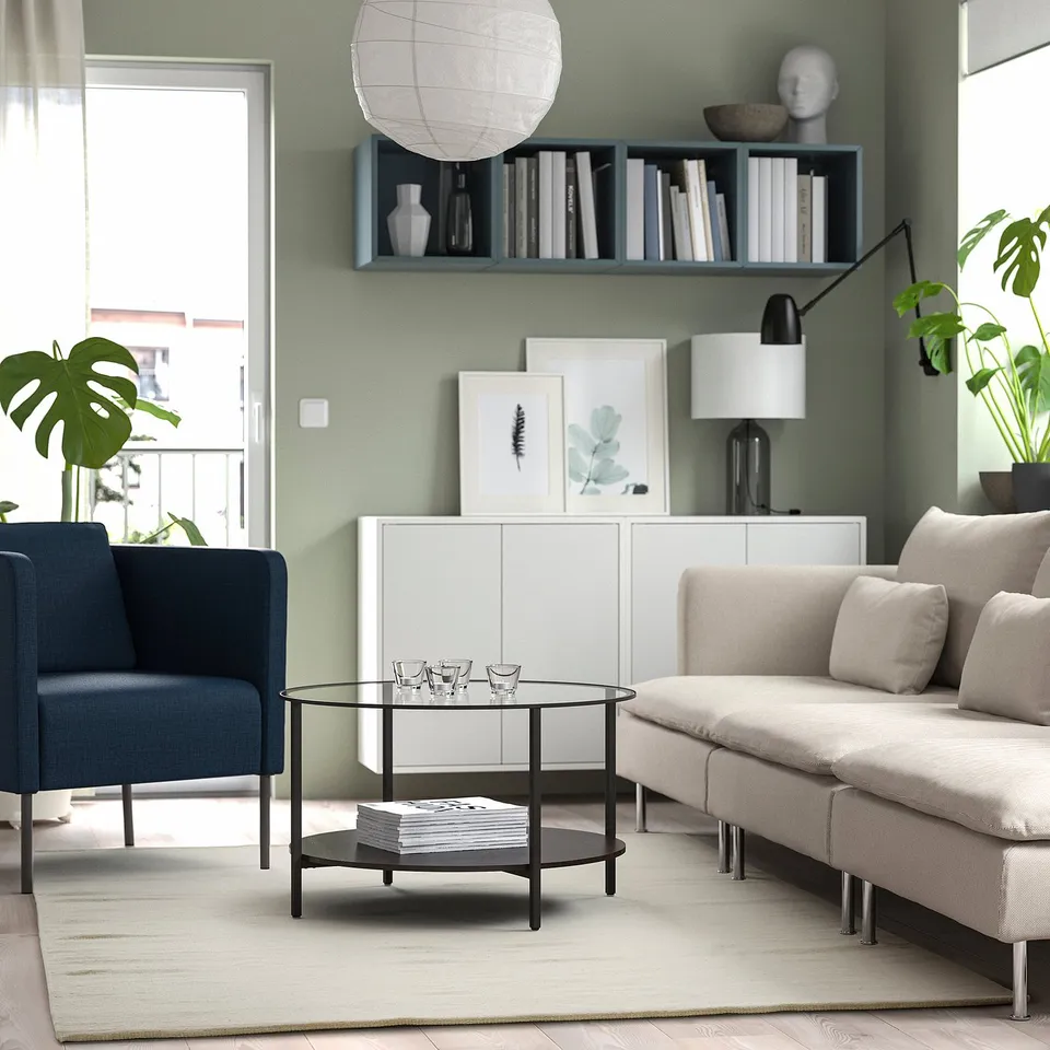 Modern living room with a light beige rug, featuring TIDTABELL coffee table, beige sofa, blue chair, glass-topped side table with books and glasses.