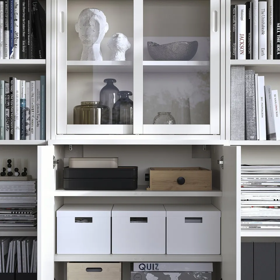 Modern white bookshelf with glass doors, displaying decorative vases and books, featuring integrated storage boxes.