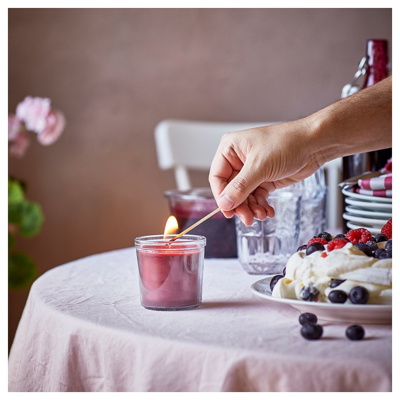 A hand lights a small, red STÖRTSKÖN candle on a table, next to a dessert, glasses, and flowers.
