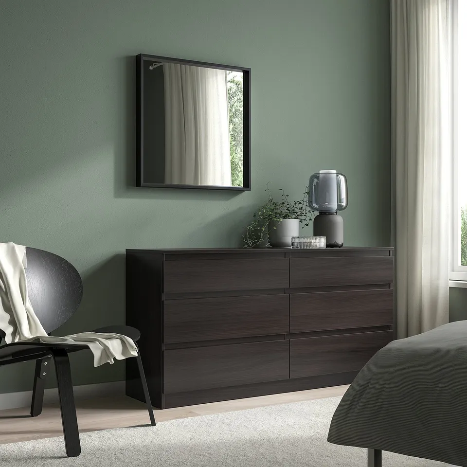 Brown 6-drawer NISSEDAL dresser with lamp, candles, plant. Black chair with cloth, bed partially seen.