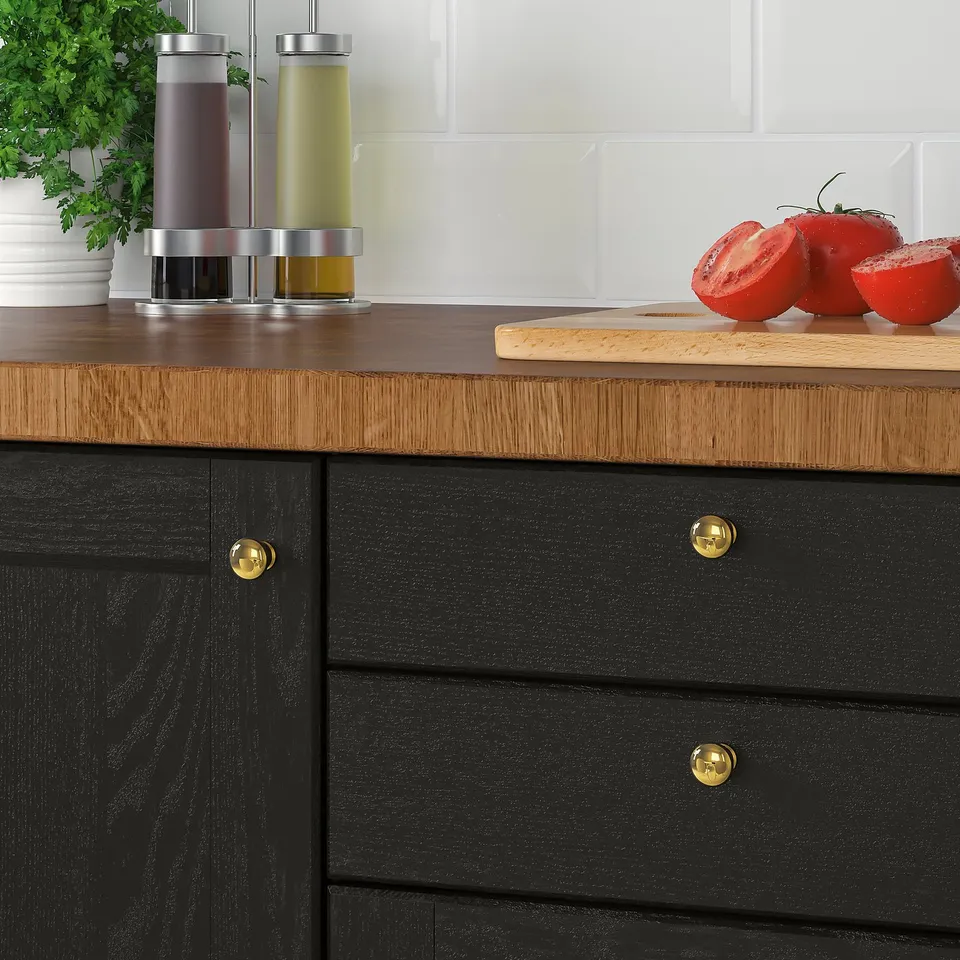 Kitchen counter with SKOGSÅ wood top, black cabinet, gold knobs, tomato, knife, oil bottles, plant. 25-year guarantee, durable, sandable.