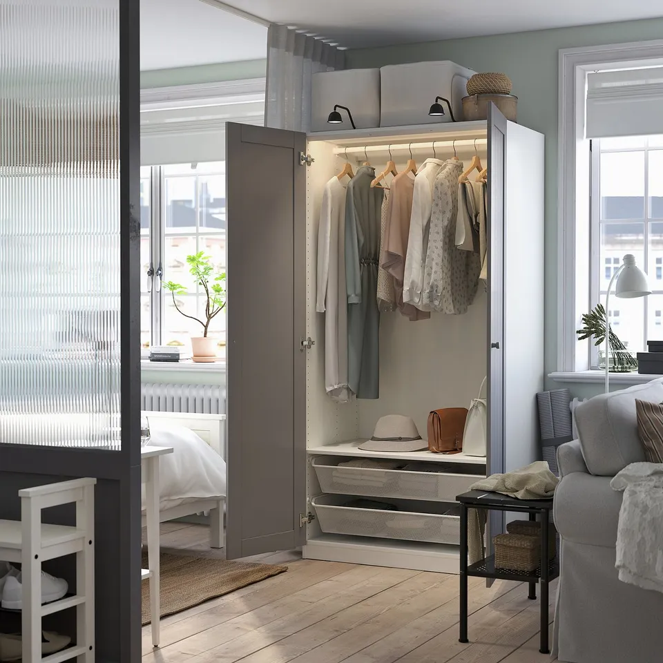 Modern bedroom with open wardrobe showing clothes, baskets, and lighting.