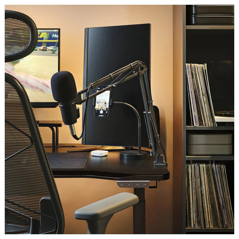 Gaming desk with black mesh chair, monitor, and microphone setup for streaming or recording. Desk has a black surface and metal base.