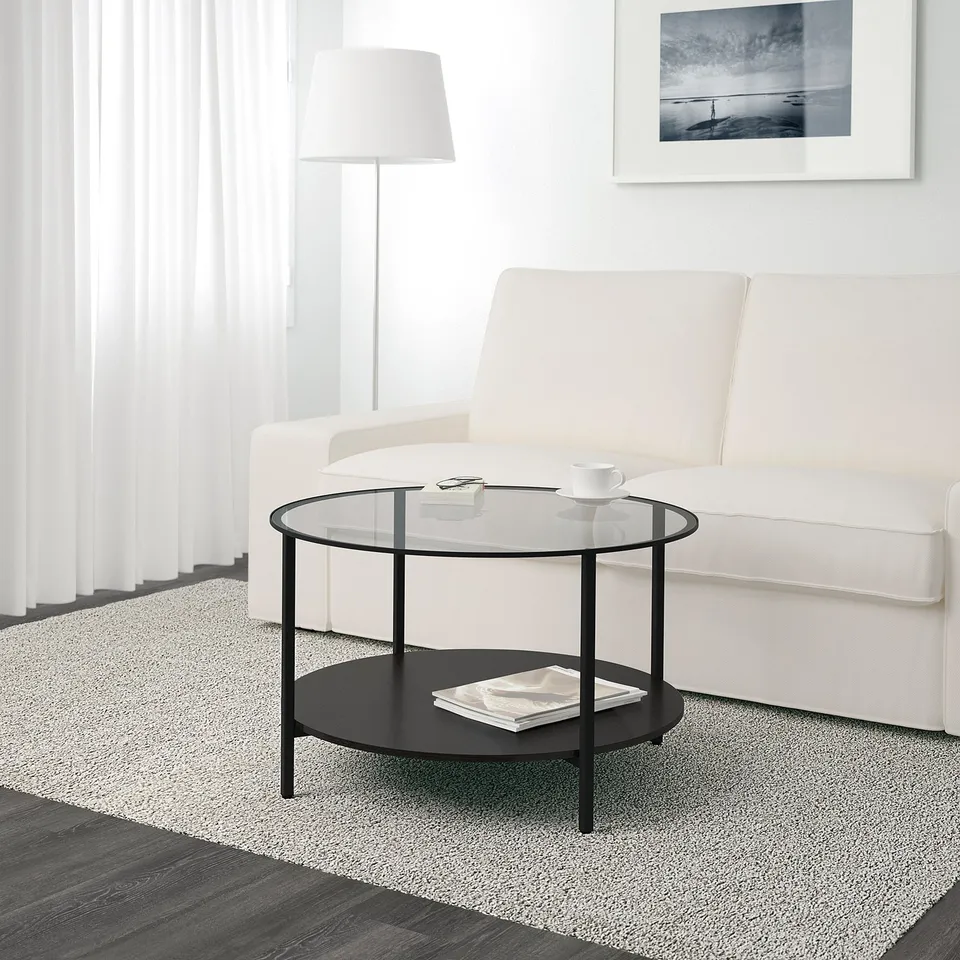 Minimalist glass coffee table with metal legs. Holds coffee mug and magazines; sits before white sofa.