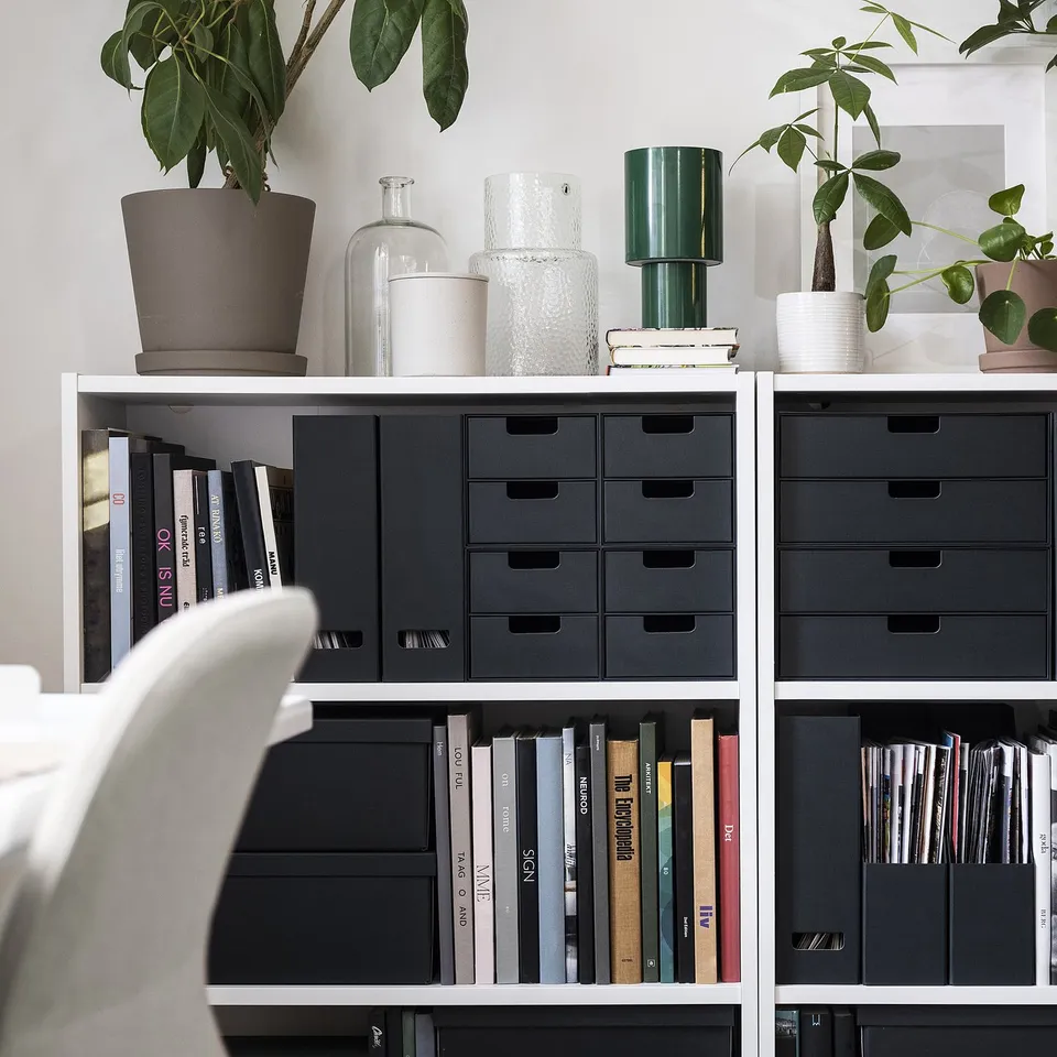 Black FJÄDERHARV boxes stored in white shelves, holding books and devices, with plants and vases on top, creating a neat workspace.