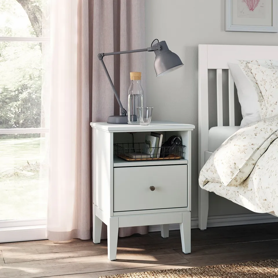 Bedroom scene: white IDANÄS bedside table, grey lamp, glass carafe, wooden basket with books, bedside table by window with curtains.