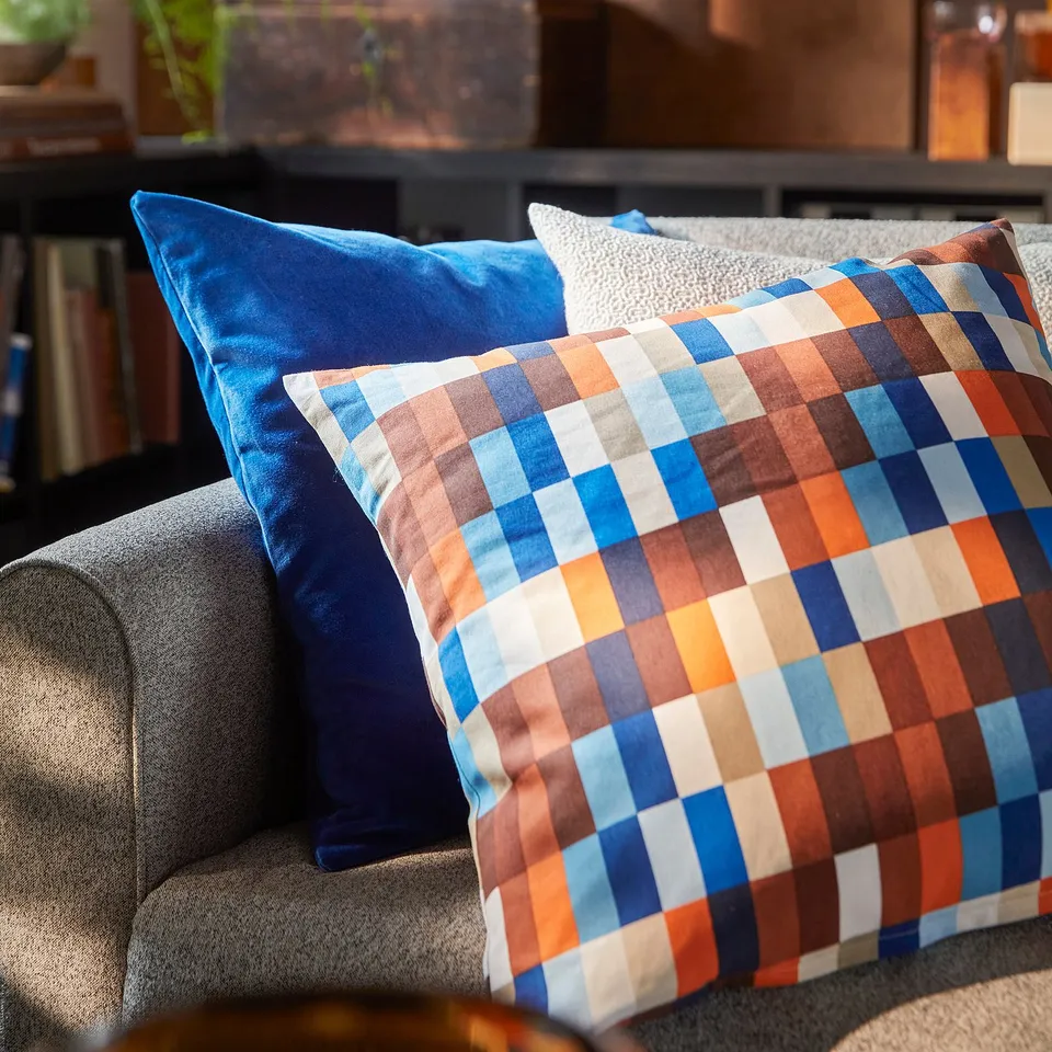 Brightly coloured chequered LÖVNATURSKOG cushion on grey couch, near blue pillow and dark shelf with books.