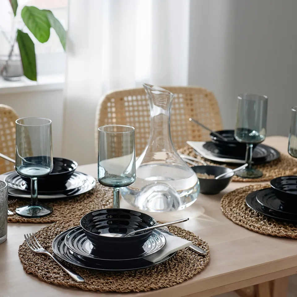 Elegant table setting with black patterned dinnerware and clear glasses on woven placemats, featuring a water pitcher.
