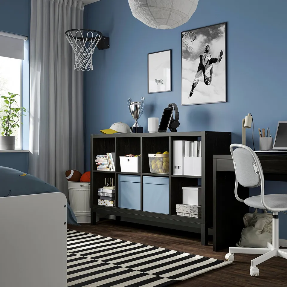 Teen bedroom with KALLAX shelf holding sports gear, trophies, books, and a basketball hoop. Striped rug and white chair are nearby.