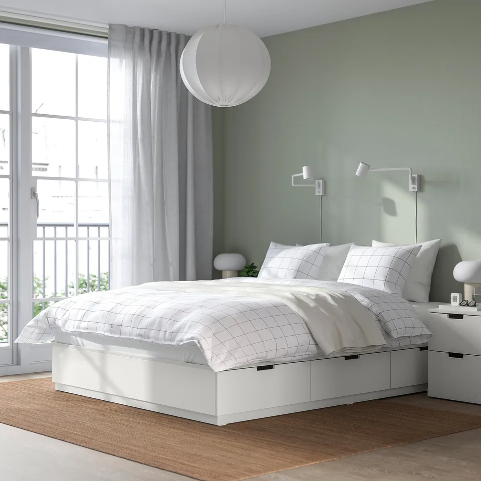 Modern minimalist bedroom: white NORDLI bed with storage drawers, grid bedding, wall lamps, large window with curtains.
