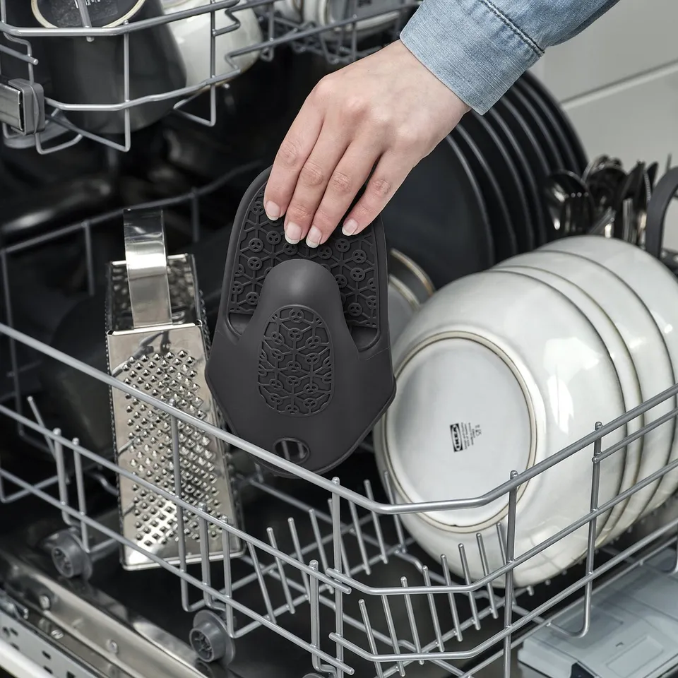 Hand placing black SANDVIVA silicone trivet on dishwasher rack, protecting fingers and helping secure items.