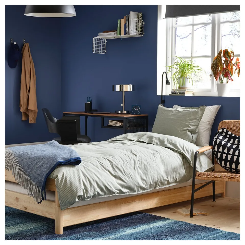 Modern blue bedroom with wooden bed, grey bedding, a desk, and plants.