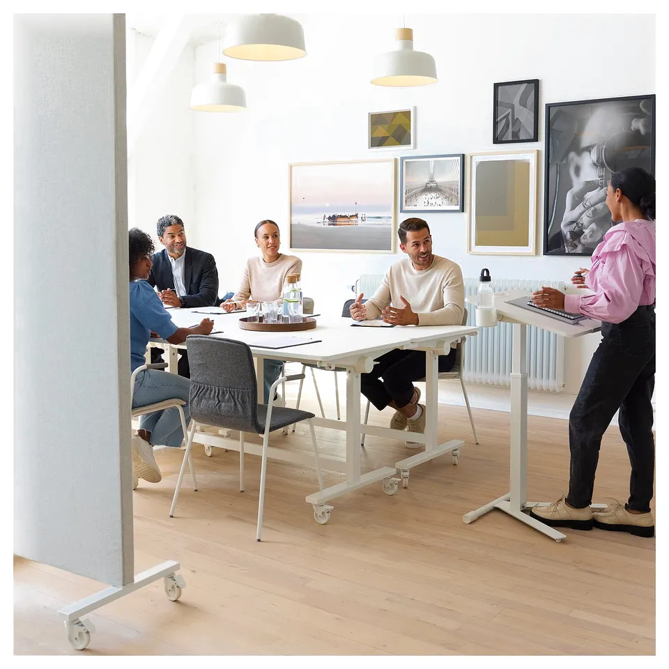 Office scene: four seated, one standing at adjustable MITTZON table. Modern design with lights and art.