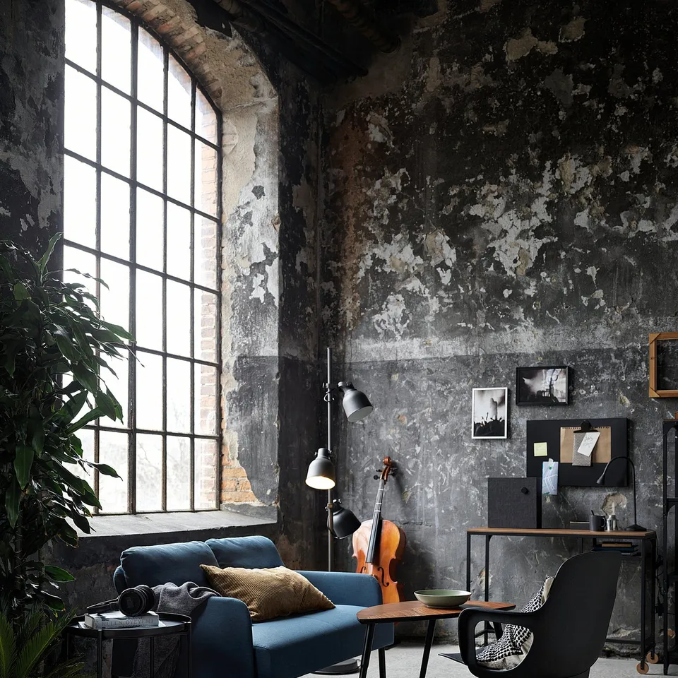 Industrial style living room with blue GLOSTAD sofa, modern desk, black chair, large window, plants, and rustic walls.
