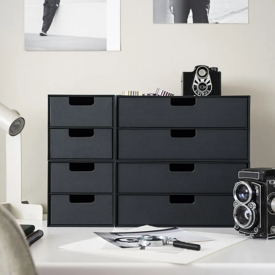 Modern black desk with stacking FJÄDERHARV mini chests, small vintage camera, and scattered papers. Minimalist, efficient storage solution.