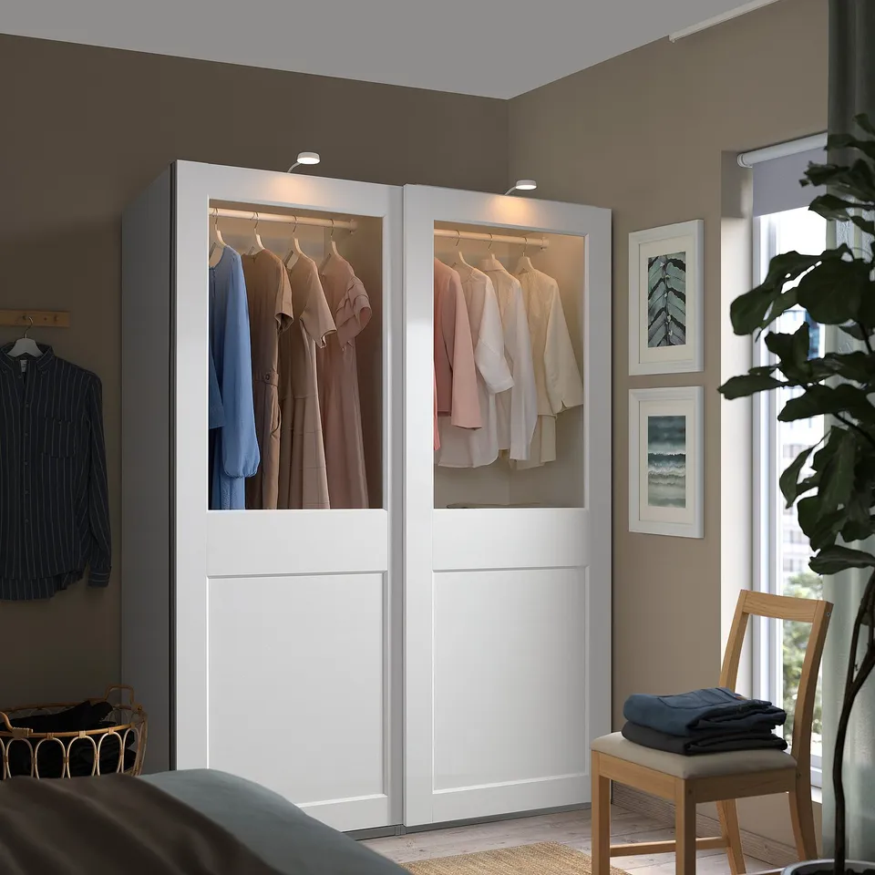 Modern white wardrobe with glass doors, clothes inside, beside window with pictures. Nearby chair has folded clothes, plant adds greenery.