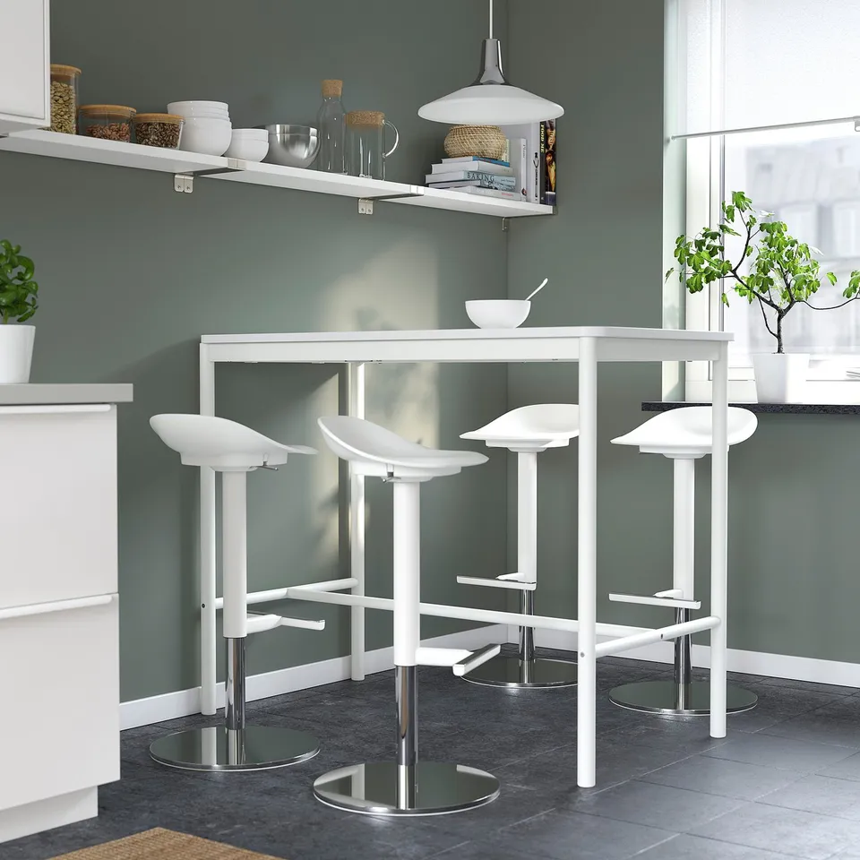Modern kitchen with white bar stools. Four minimalist chairs surround a compact table under a shelf, against green walls and grey tiles.