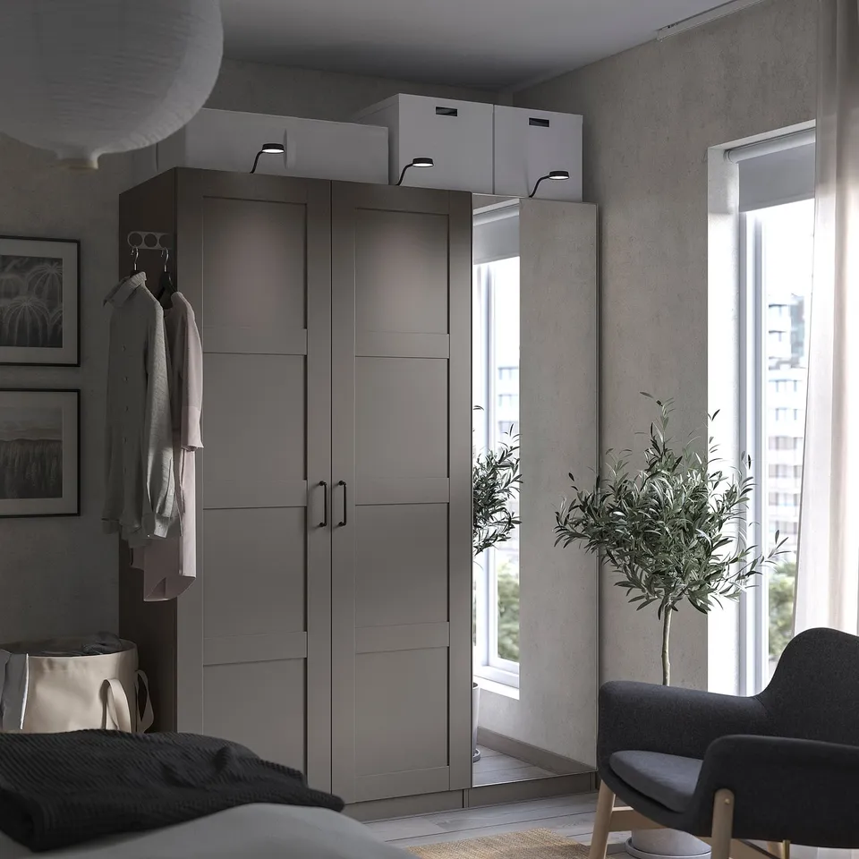Modern bedroom with grey PAX wardrobe, mirrored doors, hanging clothes, white boxes, plant, and grey chair by window.