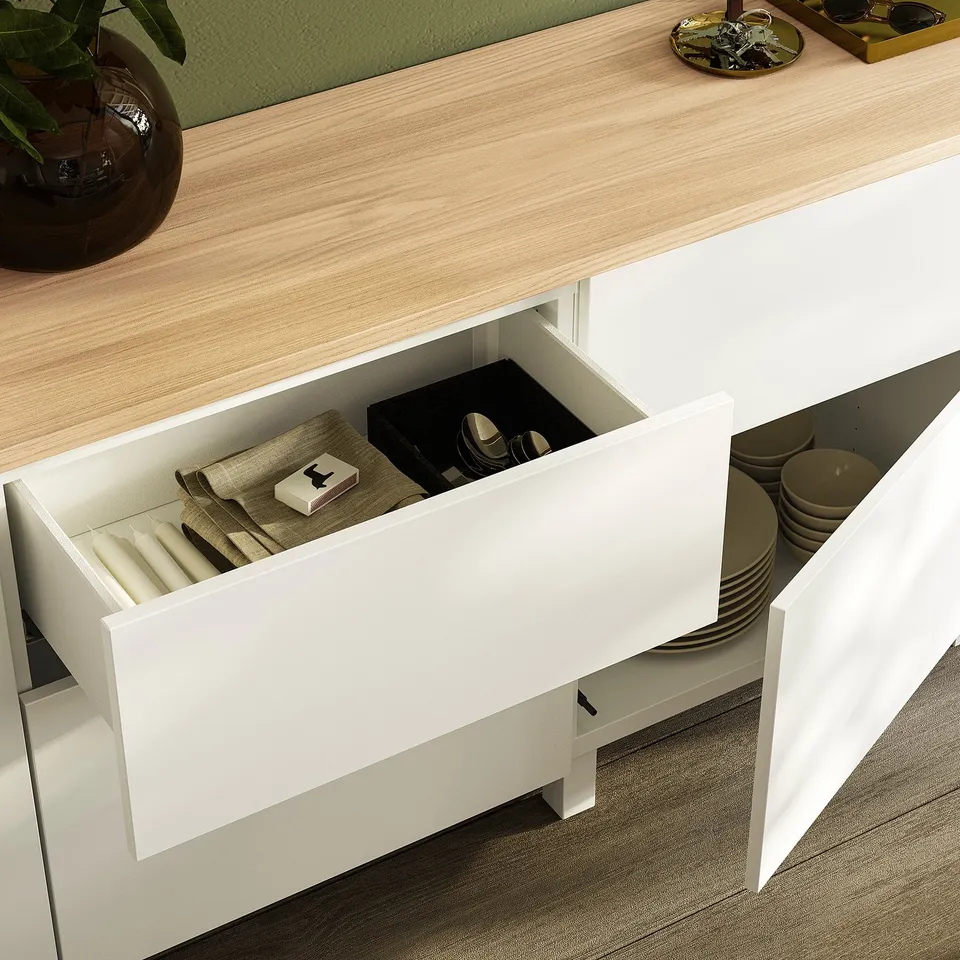 Open white kitchen drawer showing cutlery, napkins, and matchbox, with wooden countertop and plant.