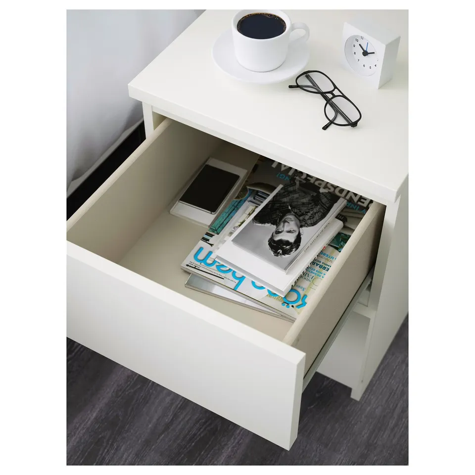 A white bedside table with an open drawer showing magazines and a phone. On top, theres a cup of coffee, glasses, and a clock.