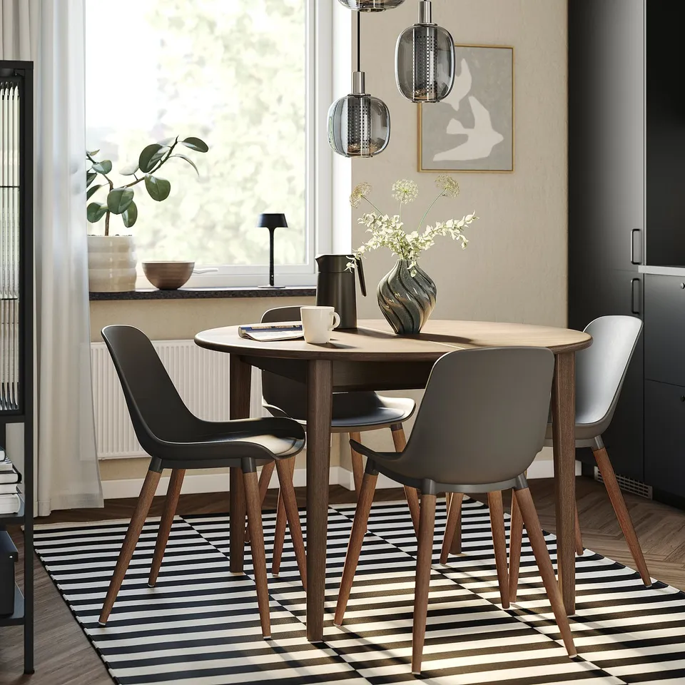 Modern dining table SKANSNÄS, small, round, wooden with 4 dark grey chairs. Under soft pendant lights, near large window.