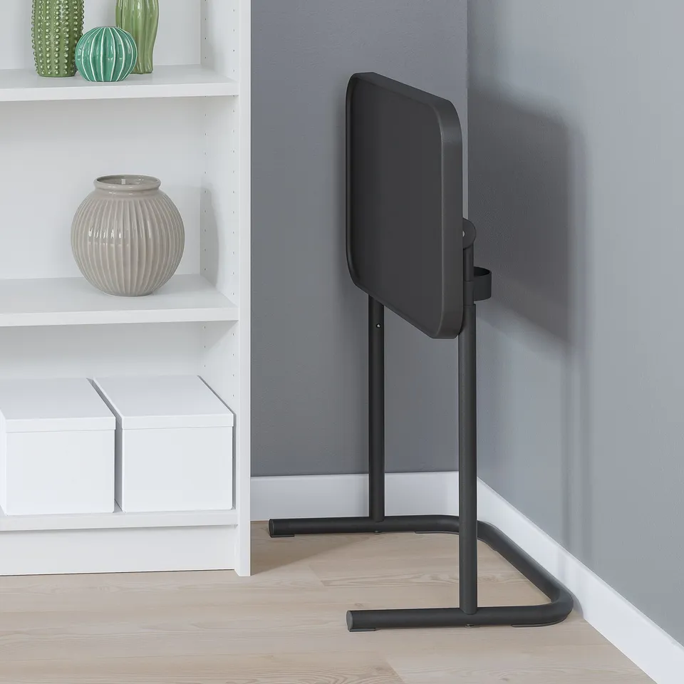 Black laptop stand in room, adjustable height, tilted top, next to shelves.
