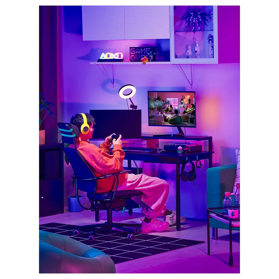 In a neon-lit room, a person plays a video game at a height-adjustable desk with a monitor, keyboard, and yellow headphones.