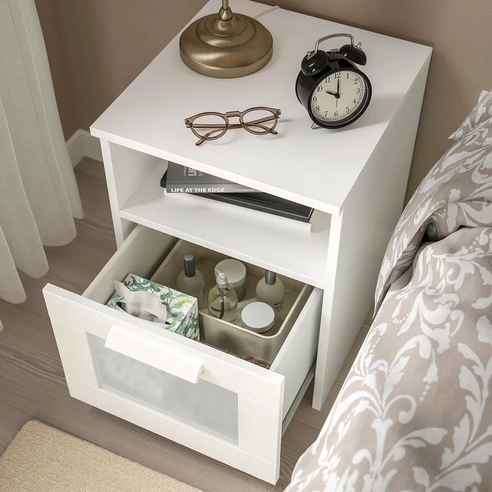 White bedside table with open drawer showing bottles, next to bed with clock and lamp on top.